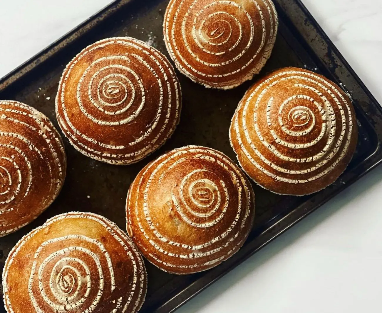Six round sourdough boules with spiral flour scoring on a baking tray
